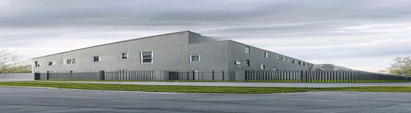 Panoramic view of a long modern single-storey institutional building with grey concrete walls and narrow vertical windows, a high perimeter fence and a closed steel gate in the foreground, soft overcast light over a tarmac forecourt