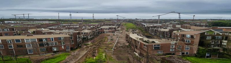 Panoramic aerial view of a half-built Irish housing estate in a green-field margin outside Dublin, rows of brick and block two-storey semi-detached houses in various stages of construction, scaffolding on some, bare foundations on others, cranes in the distance, grey overcast Irish sky