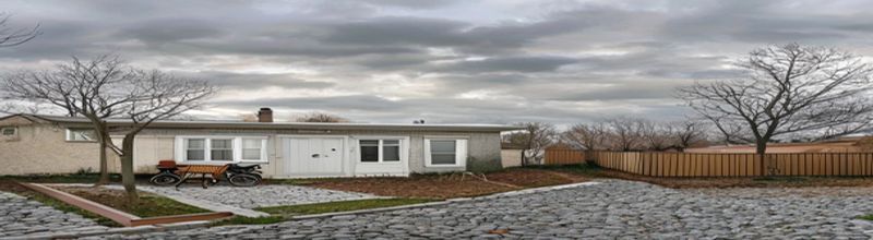 Panoramic view of a single-storey Irish residential disability-service bungalow exterior in late afternoon, pebbledash walls with a wheelchair-accessible ramp leading to a front door, a small fenced garden with a wooden bench, a cloudy pale-grey sky and bare winter trees in the distance, muted domestic institutional palette