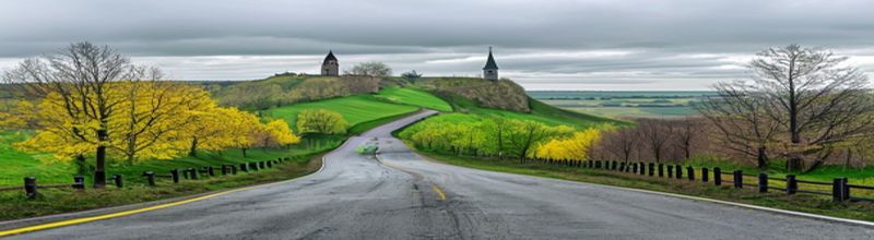 An empty rolling country road in the Belgian Ardennes climbing into low green hills under a heavy grey spring sky, leafless trees lining a metal crash barrier on the right, soft yellow-green new foliage on the verges, two distant church towers on a hill, no riders, no spectators