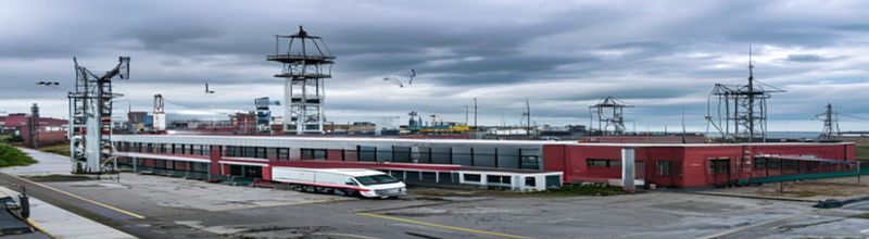Panoramic view of a red-brick industrial research park in Belfast on an overcast day, large rectangular facility with tall aluminium-clad cleanroom block, loading bay with a white unmarked van parked, a radar dome on the roof and telecommunications mast, cranes and shipyard gantries visible in the distant Belfast harbour, wet tarmac, green and grey tones