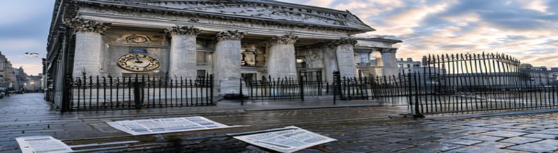 Panoramic view of the Bank of England&rsquo;s neoclassical facade on Threadneedle Street in early morning light, grey limestone columns and iron gates, ornate clock face visible, wet cobblestones reflecting the sky, scattered printed financial stability report on a wrought-iron bench in the foreground, no people no faces no hands