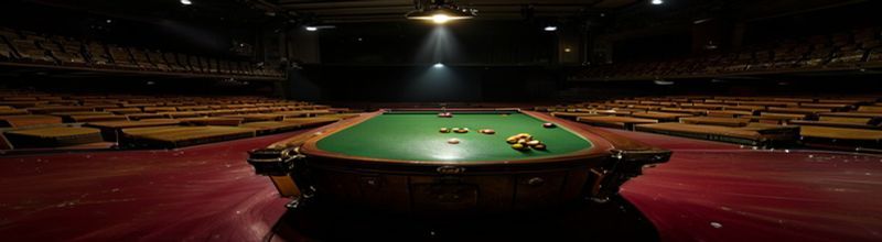 A panoramic interior view of the Crucible Theatre in Sheffield, the green baize snooker table lit by a single overhead lamp at the centre of an otherwise dark thrust-stage arena, polished red and yellow balls racked, ranks of empty steeply tiered seats receding into shadow on three sides, no players no spectators no people no faces no hands