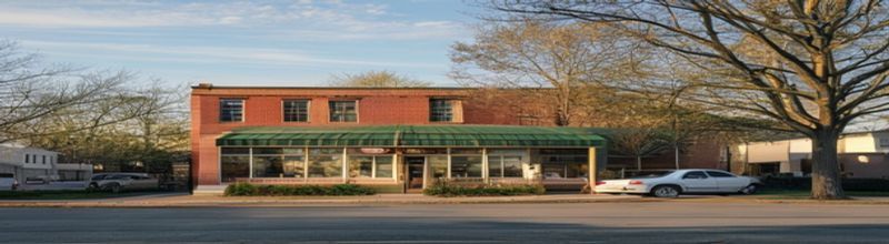 Panoramic view of a small Midwestern community bank branch on a quiet town main street, two-storey red-brick building with a green awning and gold lettering above the door, an empty parking lot with a single white pickup truck, pale morning sun glancing off the window glass, maple trees in first leaf, no people no faces no hands