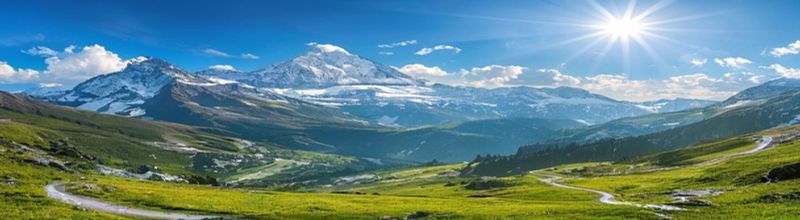 Panoramic view of the Vaud Alps under bright morning sun, the snow-capped Dents du Midi range across the horizon, a switchbacked mountain road climbing up through alpine pasture in the foreground, a Tour de Romandie banner stretched across the road in the middle distance, no people no faces no hands