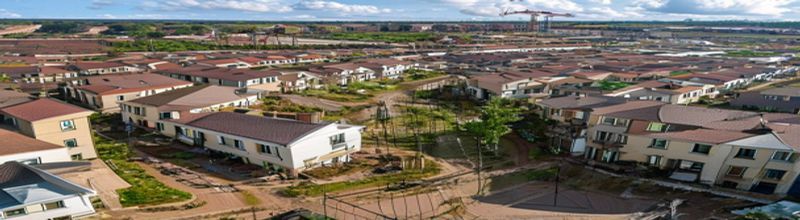 Panoramic view of a partially completed Irish suburban housing estate at dusk, neat rows of two-storey houses with grey slate roofs and pale rendered walls, scaffolding around a few unfinished units, mature hedgerows in the foreground, no people no faces no hands