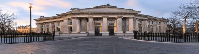 Panoramic exterior view of Leinster House Dublin at golden hour, neoclassical Georgian sandstone facade with central pediment and columns, ornate iron railings along the front, deep grey paving and bare flagpoles, soft amber light raking across the stone, no people no faces no hands