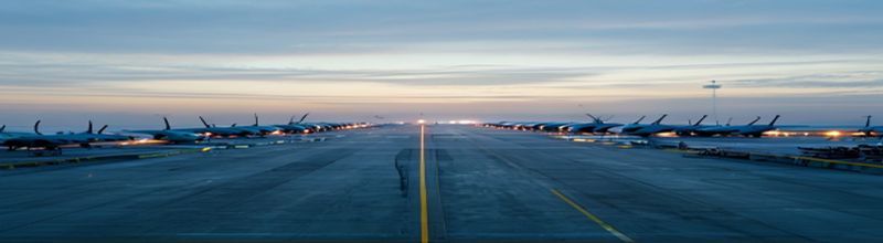 Panoramic dawn view of an empty airport apron with a long row of identical narrow-body jets parked at jet bridges, fuel bowsers lined up at the end of the row, soft pale-blue runway lights still illuminated, a control tower silhouetted against a colour-graded sky, no people no faces no hands