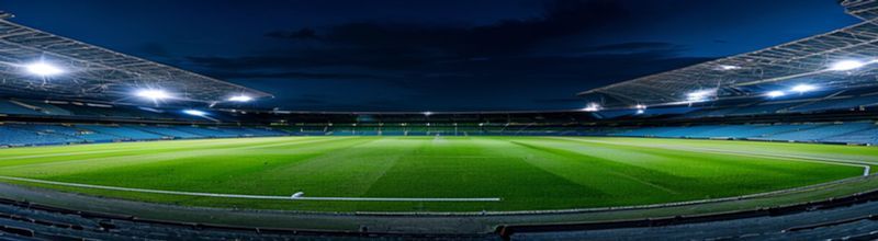 Panoramic view of an empty European football stadium at night, the pitch brightly lit by tall floodlights, the dark blue sky just above the rim of the upper tier, two banks of empty grandstand seating either side of the centre line, no people no faces no hands
