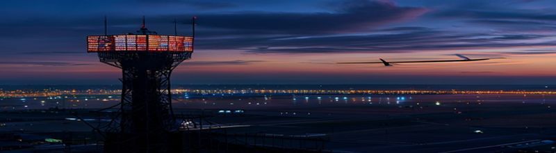 Panoramic view of an air traffic control tower at a regional airport at dusk, dark glass cab glowing with screens at the top, runway lights stretching into the distance, dark sky with the silhouette of a parked aircraft on the apron, no people no faces no hands