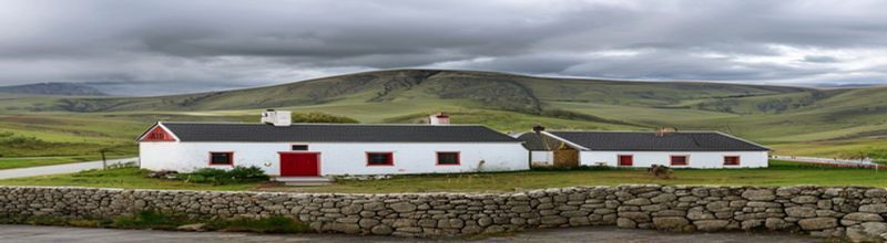 Panoramic view of a small village community hall in rural County Down, white-painted stone building with a slate roof and red front door, a low stone wall in the foreground, rolling green Mourne Mountain hills in the background under a soft cloudy sky, no people no faces no hands