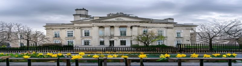 Panoramic wide view of Leinster House facade in Dublin at early morning with grey overcast sky, the limestone classical front and central pediment in sharp focus, Merrion Square railings in foreground with daffodils in flower beds, a few empty wooden benches, soft diffused light, no people no faces no hands