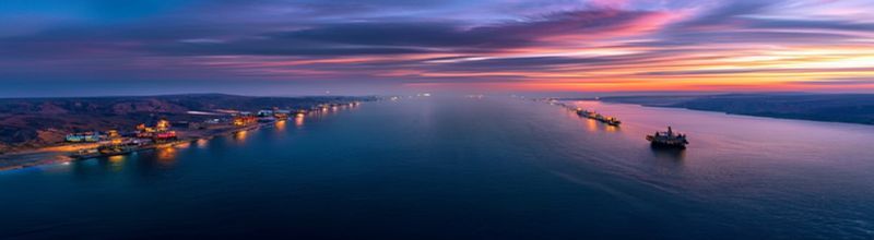 Panoramic view of the Strait of Hormuz at dusk, narrow shipping lane between rugged coastlines, oil tankers under way in single file, calm sea, distant haze, no people no faces no hands