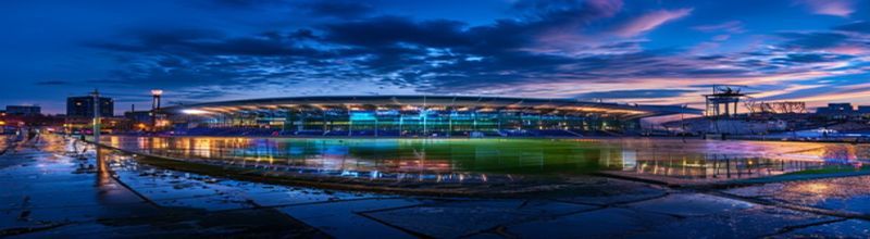 Panoramic view of an empty European football stadium at night, the pitch brightly lit by tall floodlights, the dark blue sky just above the rim of the upper tier, two banks of empty grandstand seating either side of the centre line, no people no faces no hands