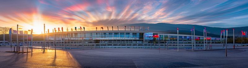 Panoramic view of the World Health Organization headquarters in Geneva at sunrise with mountains in the background, the curved modernist building reflecting morning light, member-state flags on tall poles in a row in the foreground courtyard, no people no faces no hands