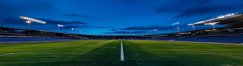 Panoramic view of an empty European football stadium at night, the pitch brightly lit by tall floodlights, the dark blue sky just above the rim of the upper tier, two banks of empty grandstand seating either side of the centre line, no people no faces no hands