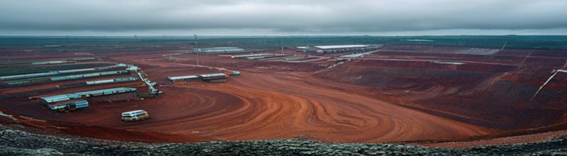 Panoramic wide-angle photograph of an open-pit cobalt mine, terraced red-brown earth, haul trucks parked at the rim, metal processing sheds in the distance, overcast equatorial light, documentary realism, no people no faces no hands