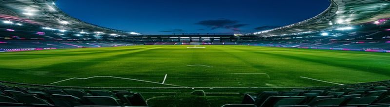 Panoramic view of a Premier League stadium at twilight from the upper tier, empty floodlit pitch, white painted markings vivid against deep green grass, illuminated stand seats in club colours, scoreboard glow, no people no faces no hands