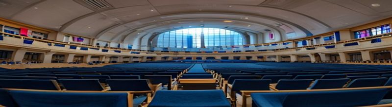 Panoramic interior of the European Parliament hemicycle in Brussels, empty blue committee seats arched in tiers, EU and member state flags hung along the back wall, soft daylight from skylight, no people no faces no hands