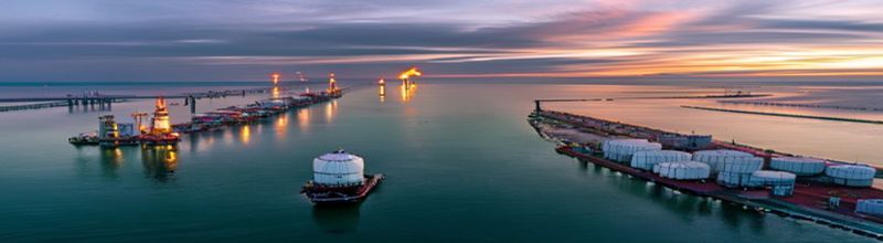 Panoramic dawn aerial view of a vast oil export terminal on the Texas Gulf Coast with rows of cylindrical white storage tanks reflecting first light, a long jetty extending into calm water with two anchored tankers riding low in the water, dredged channel marked by red and green buoys, distant flare stack burning a thin orange flame against pale grey sky, no people no faces no hands
