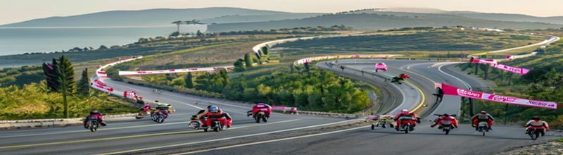 Panoramic wide-angle view of a Giro d'Italia race convoy on an Italian coastal road in soft early-morning light, a tight peloton of cyclists in colourful kit stretched along a curve of asphalt above the Adriatic, white official cars and red Vespa motorbikes following, pink Giro logo banners on roadside barriers, distant olive groves on the hillside, sharp natural detail on the road surface and barriers, no people no faces no hands