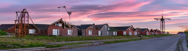 Panoramic view of a new Irish housing estate at the edge of a green field at dawn, three rows of red-brick semi-detached houses under construction with timber roof trusses exposed on the nearest row, a small tower crane silhouetted against a pink and grey sky, neat tarmac kerb stones marking the new estate road, low stone wall on the field boundary, no people no faces no hands