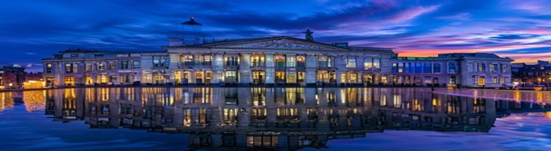 Panoramic view of the Treasury Building on Grand Canal Street in Dublin at dusk, modern glass and limestone facade reflecting the surrounding canal water, an Irish tricolour above the entrance under a deep blue evening sky, no people no faces no hands