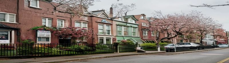 Panoramic view of a row of red-brick Edwardian terraced houses along a Dublin residential street at midday, painted front doors in deep greens and reds, neat front gardens with low railings, a cherry tree in mid-blossom, a printed for-sale board on one gable, parked cars along the kerb, pale overcast spring light, no people no faces no hands