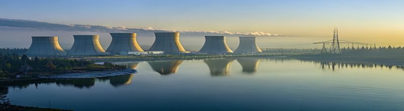 Panoramic view of a large nuclear power station's cooling-towers complex at dawn from across a wide reservoir, six identical hyperbolic cooling towers in two rows beside reactor buildings, a high-voltage transmission gantry with multiple lines running off to the right, no people no faces no hands
