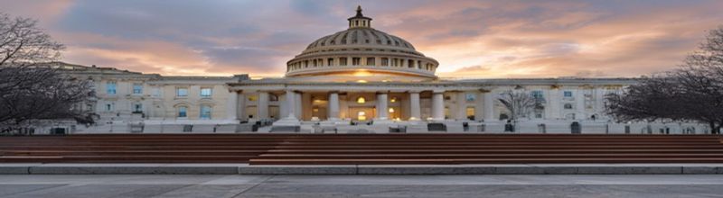 Panoramic exterior view of the United States Capitol's Senate wing at dawn, the dome partially lit by low rose-gold light against an overcast sky, the broad east-front steps in the foreground with a single empty bench, no people no faces no hands