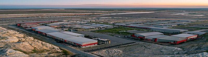 Panoramic aerial view of a U.S. Army industrial ammunition plant on a flat midwestern site at dawn, rows of grey corrugated production sheds and red-brick administrative buildings inside a perimeter fence, a small rail spur with covered wagons, exposed bedrock and a quarry pit cut into the grassland behind the plant showing pale grey limestone strata, no people no faces no hands