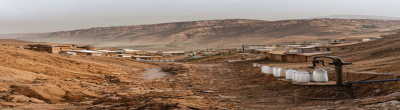 Panoramic view of a dry rocky valley in western Afghanistan at midday under a hazy pale sky, brown clay-walled village houses on a hillside, an empty earthen irrigation channel cut along the slope, distant high mountain ridge under a thin layer of dust, a row of empty white plastic water jerry-cans stacked beside a hand pump, no people no faces no hands